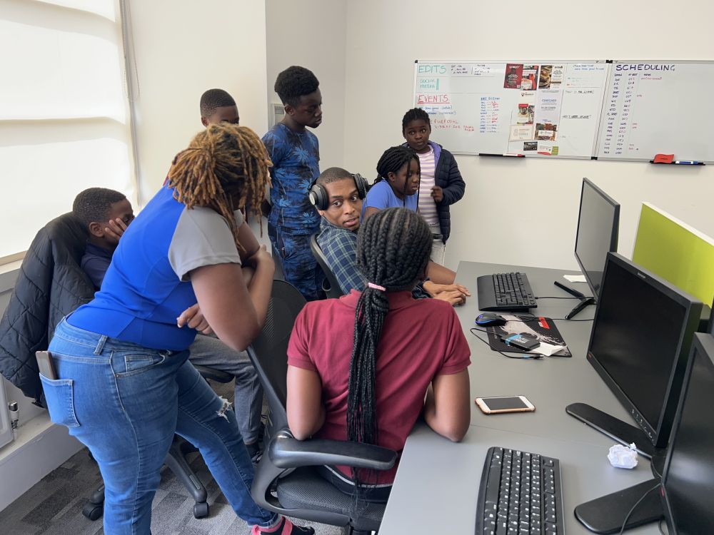 8 young people gathered around a computer who appear to be editing a podcast.